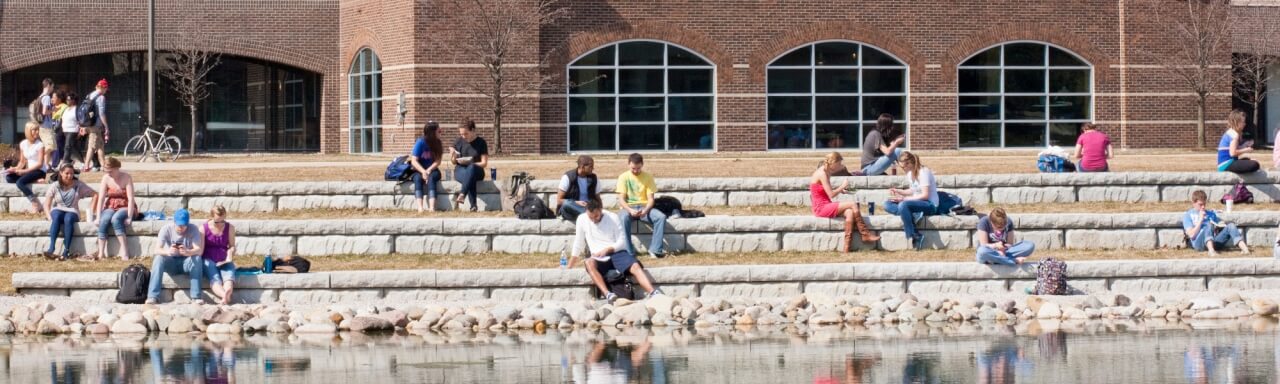 Zumberg pond featuring Kirkhof center in the background and GVSU students sitting near the pond
